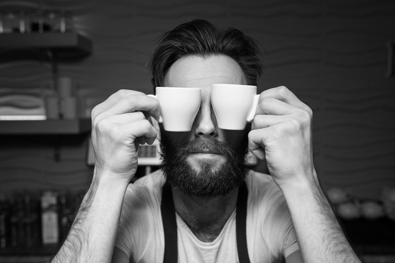 Black and white photo of a barista holding two espresso mugs over his eyes